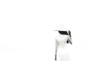 Woman in white ao dai and conical hat against a pale background